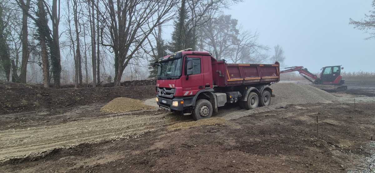 Ein roter Muldenkipper auf einer matschigen Straße in der Nähe von Bäumen und ein Bagger an einem nebligen Tag.