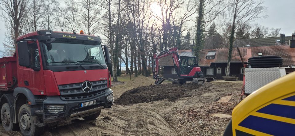 Baustelle mit einem roten Muldenkipper, einem Bagger und einem Gebäude. Erde wird bewegt.