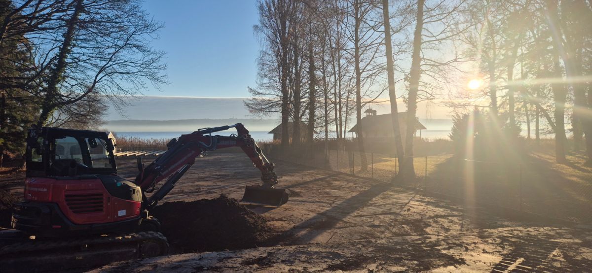 Ein kleiner roter Bagger gräbt vor einem Gewässer, durch dessen Bäume Sonnenlicht scheint.