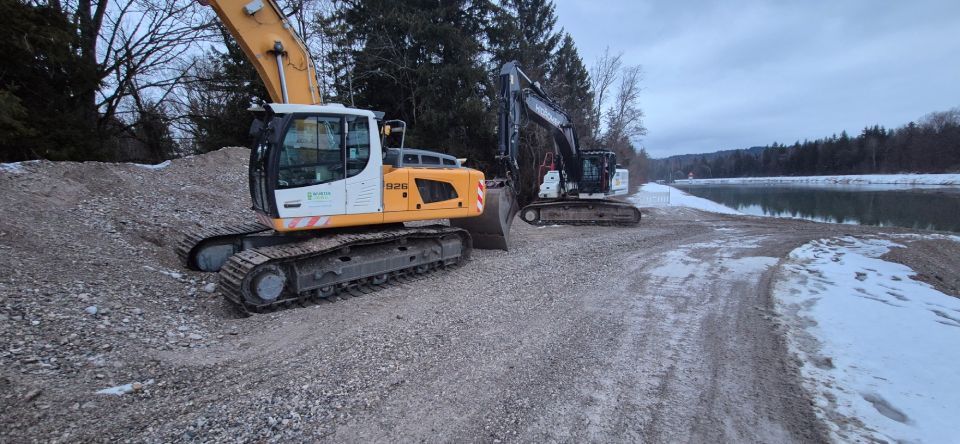 Zwei Bagger arbeiten unter bewölktem Himmel in der Nähe eines Wasserlaufs mit schneebedecktem Ufer.
