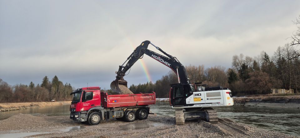 Ein Bagger belädt einen roten Muldenkipper mit Kies; bewölkter Tag, Regenbogen sichtbar.