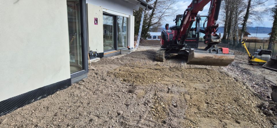 Ein Bagger gräbt Erde in der Nähe eines modernen Gebäudes mit großen Glasfenstern aus.