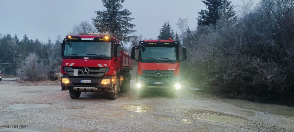 Zwei rote Mercedes-Muldenkipper auf einem Feldweg, einer davon mit eingeschalteten Scheinwerfern, in einem Waldgebiet.