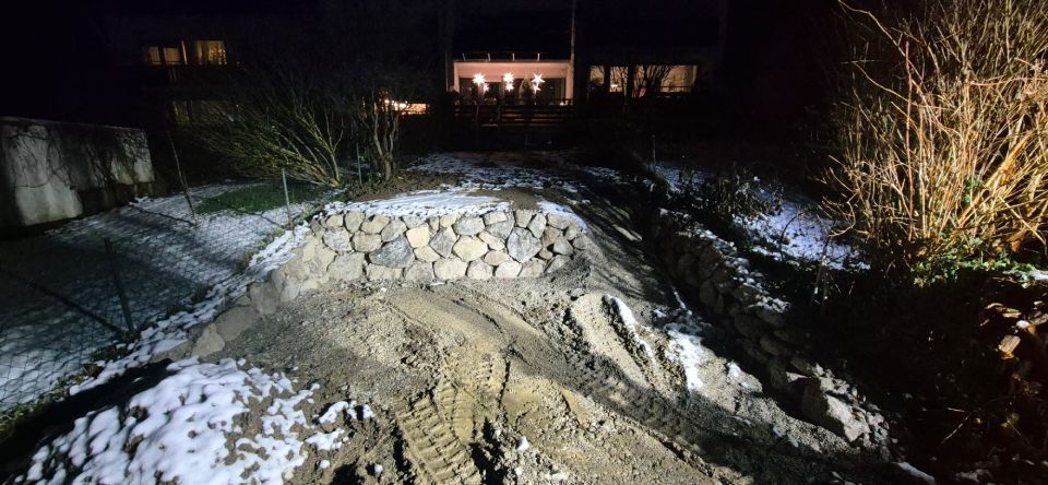 Nachtansicht eines schneebedeckten Hügels mit einer steinernen Stützmauer und einem Haus mit Lichtern in der Ferne.