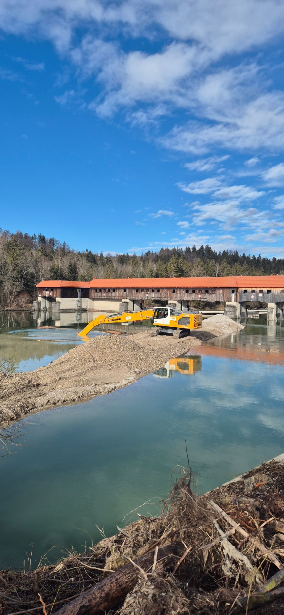 Ein gelber Bagger arbeitet an einer roten überdachten Brücke über einen Fluss. Strahlend blauer Himmel mit Wolken.