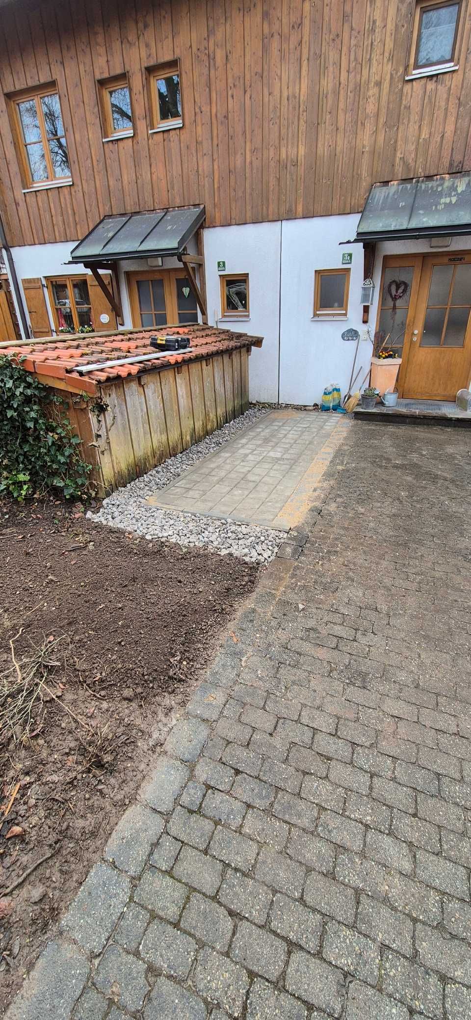 Ein Kopfsteinpflasterweg führt zu einem verwitterten Gebäude mit Holzverkleidung und kleinen Fenstern, an dessen Seite sich Erde und Kies aneinanderreihen.