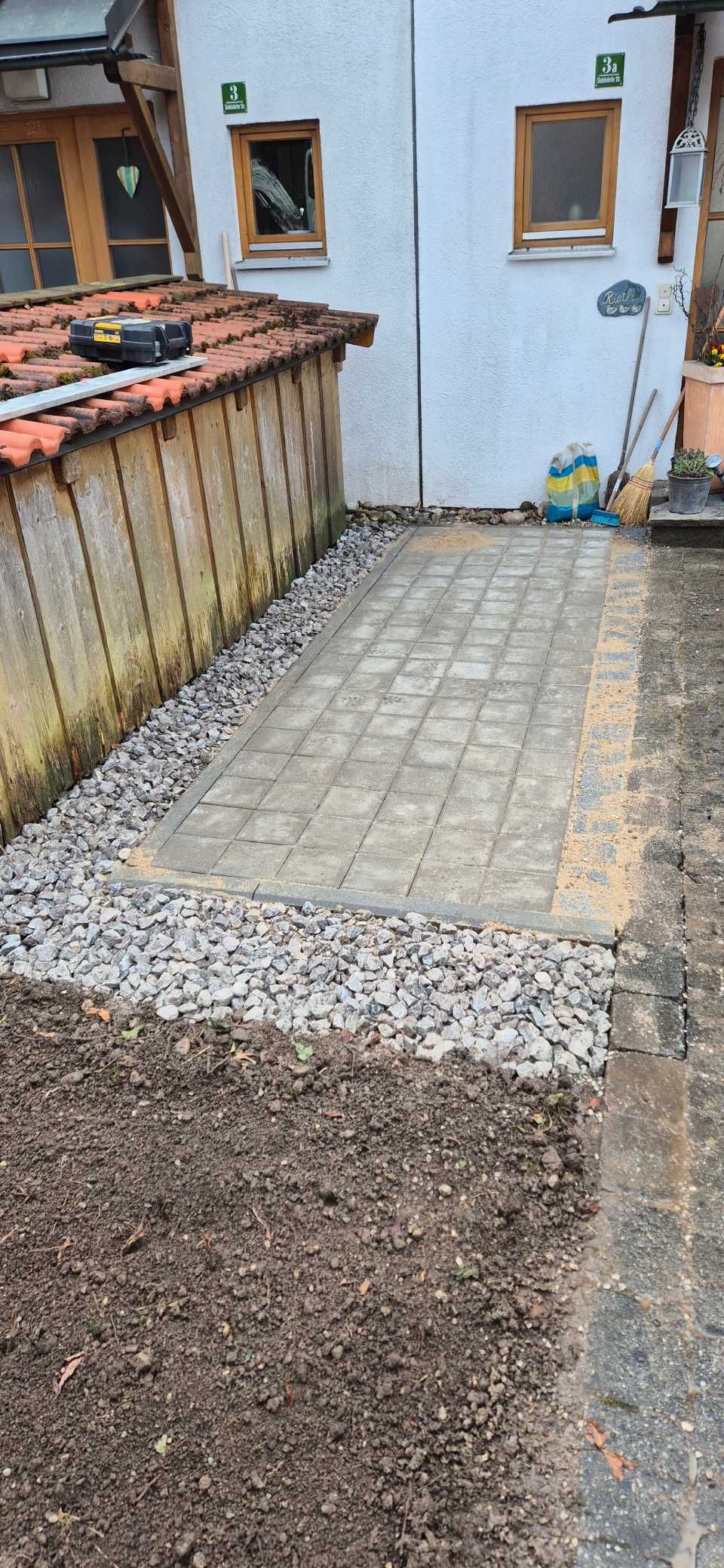 Baustelle mit Kies, Erde und einer gefliesten Fläche neben einem Gebäude mit Fenstern.