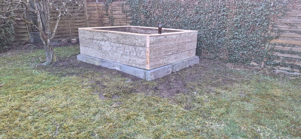 In einem Garten mit Rasen und Bäumen steht ein rechteckiges Steinbeet auf einem Betonfundament.