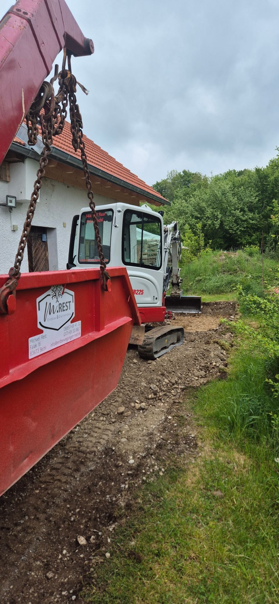 Ein roter Abrollkipper und ein Bagger in der Nähe eines weißen Gebäudes mit orangefarbenem Dach an einem bewölkten Tag.