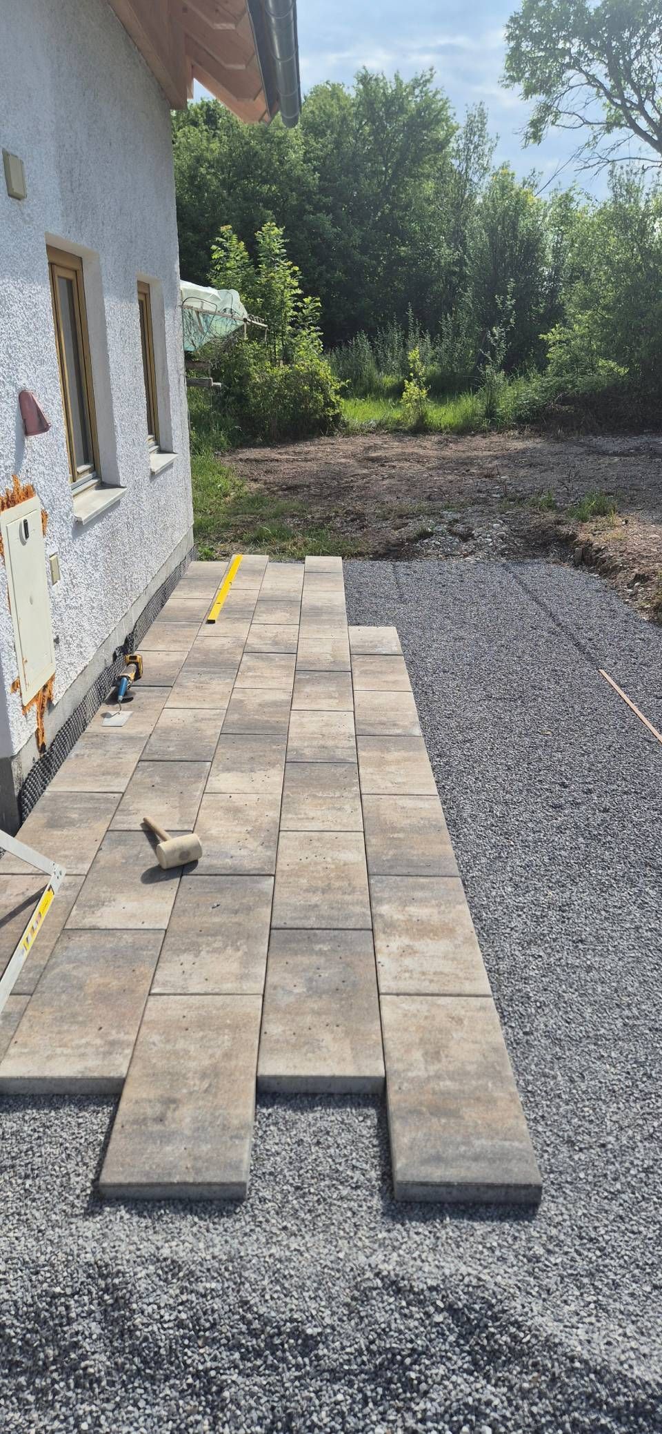 Terrassenplatten werden auf Kies neben einem Haus verlegt, im Hintergrund sind grüne Bäume zu sehen.