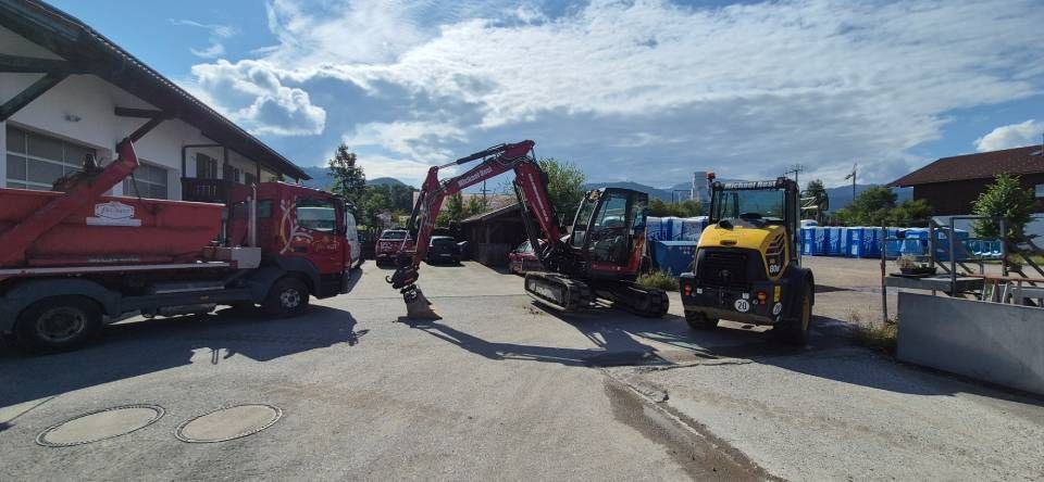 Ein roter Muldenkipper, ein Bagger und ein gelber Traktor parkten an einem sonnigen Tag vor einem Gebäude.