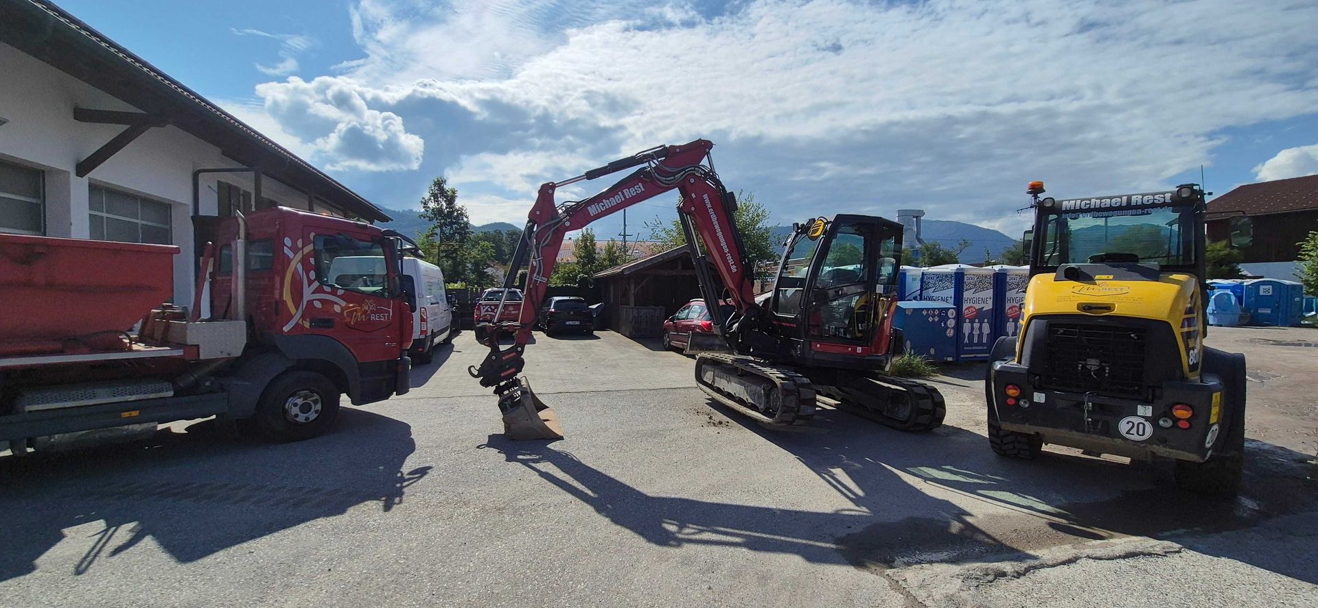 Ein roter Lastwagen, ein Bagger und ein gelbes Baufahrzeug stehen unter bewölktem Himmel vor einem Gebäude.