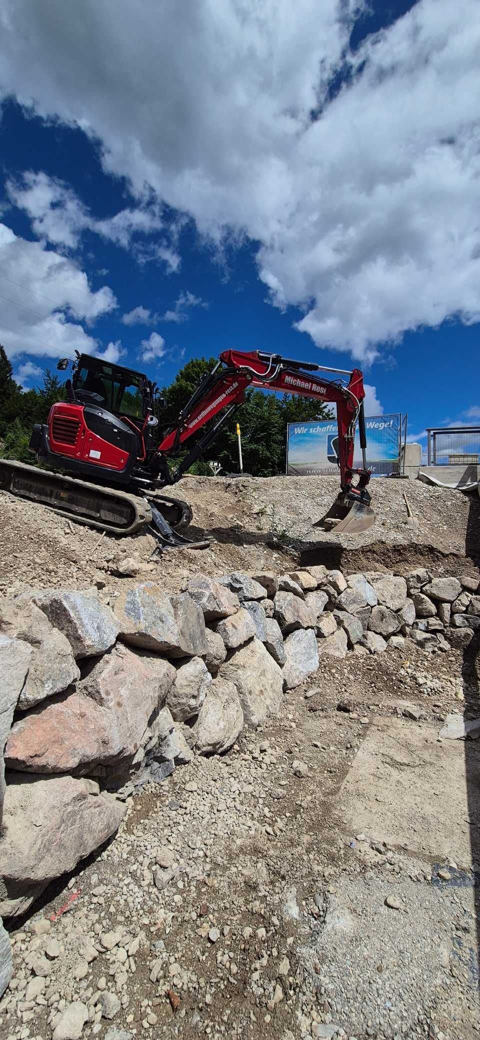 Ein roter Bagger ist auf einer Baustelle im Einsatz. Im Vordergrund sind große Felsen zu sehen, darüber ein blauer Himmel.