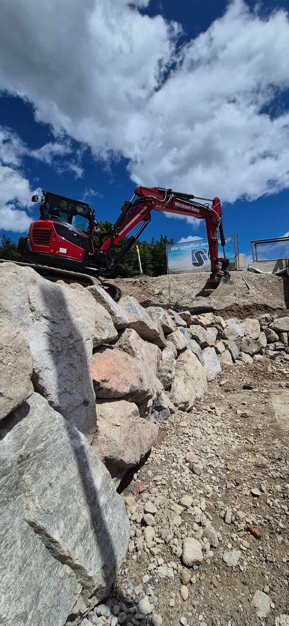 Ein roter Bagger arbeitet in einer felsigen Landschaft unter blauem Himmel mit Wolken.