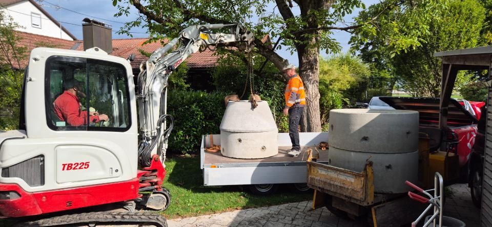 Ein Minibagger und ein Arbeiter entladen Betonbrunnenringe von einem Tieflader auf einem Hof.