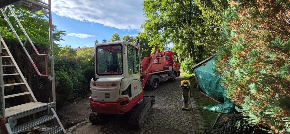 Baufahrzeuge, darunter ein kleiner Bagger, auf einer Schotterauffahrt, im Hintergrund Bäume und Baugerüste.
