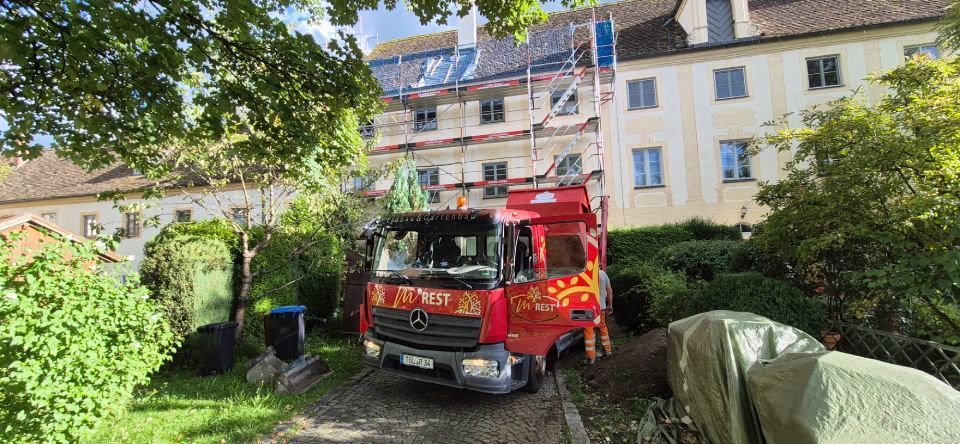Ein rotes Feuerwehrauto parkt vor einem eingerüsteten Gebäude. Grüne Bäume umgeben das Gelände.
