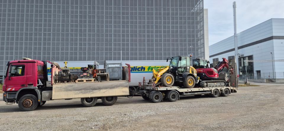 Ein roter Lkw zieht einen Anhänger mit Baumaschinen. Im Hintergrund ist ein Gebäude zu sehen.