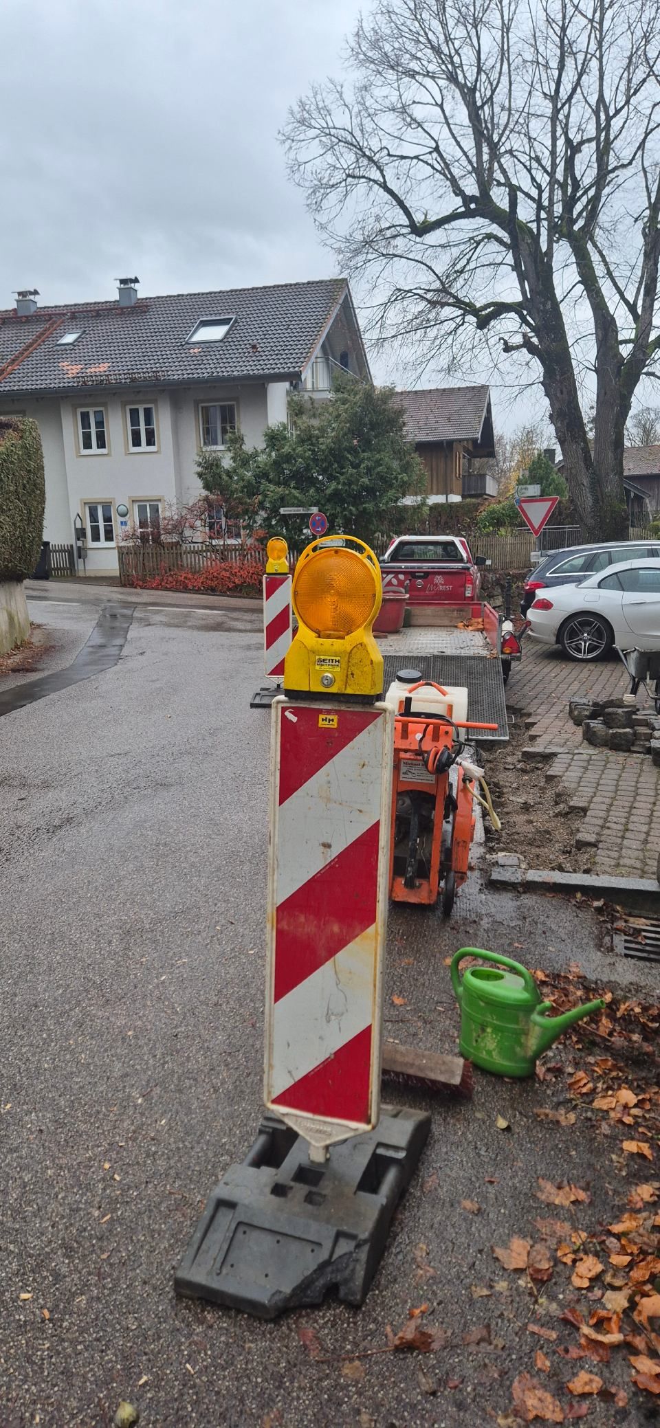 Baustelle mit rot-weiß gestreifter Absperrung und Blinklicht. Eine grüne Gießkanne liegt auf dem Boden.
