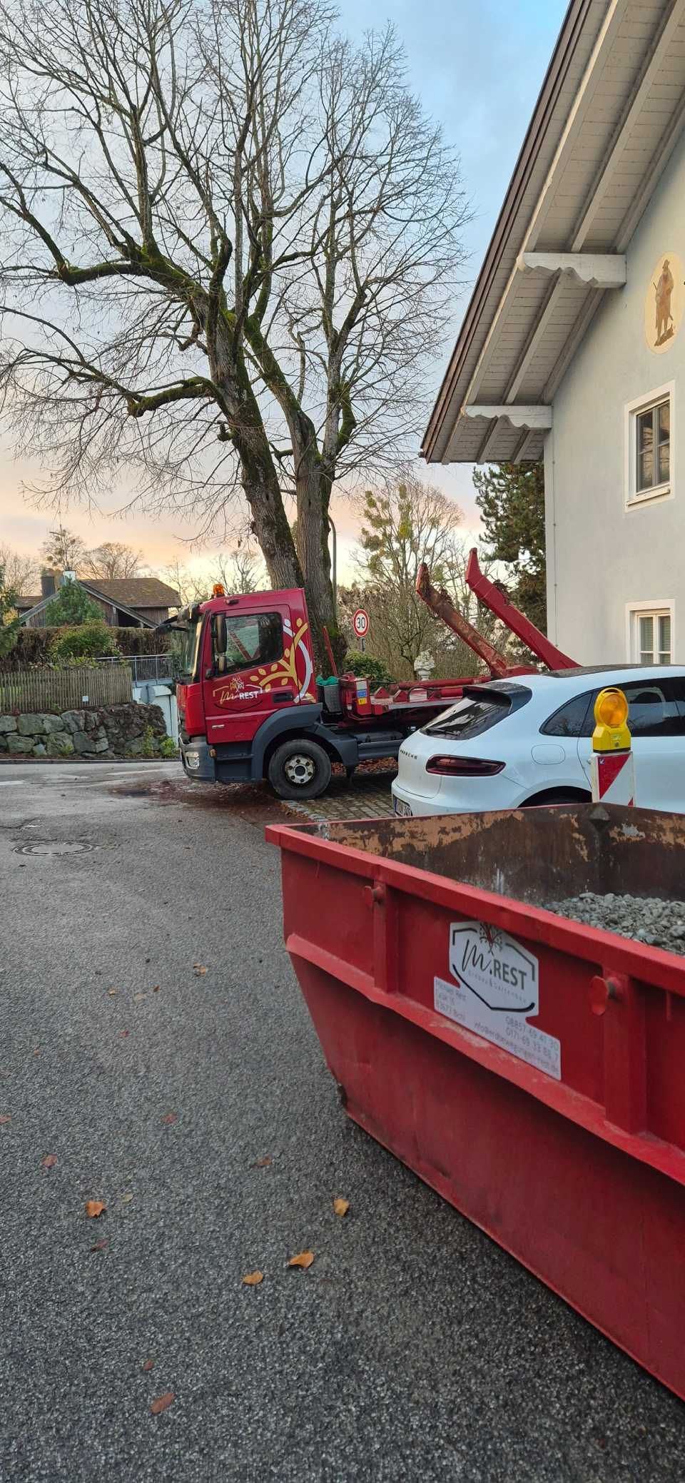 Ein roter Lastwagen, ein Baum und ein roter Müllcontainer. Ein weißes Auto und ein Gebäude sind ebenfalls zu sehen.