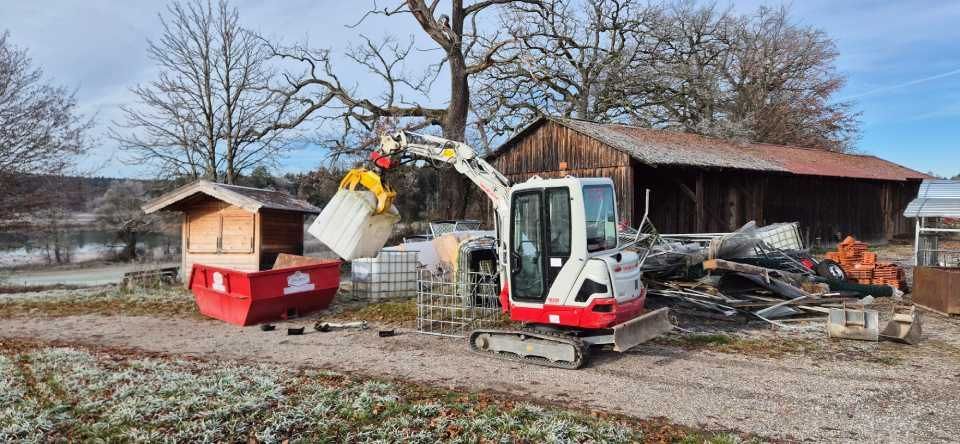 Ein Bagger belädt einen Container. Im Hintergrund sind ein kleines Holzgebäude und eine Scheune an einem frostigen Tag zu sehen.