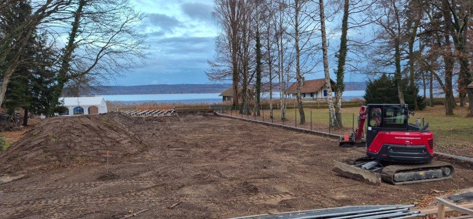 Baustelle mit einem roten Bagger in der Nähe eines Sees, Häusern und Bäumen an einem bewölkten Tag.