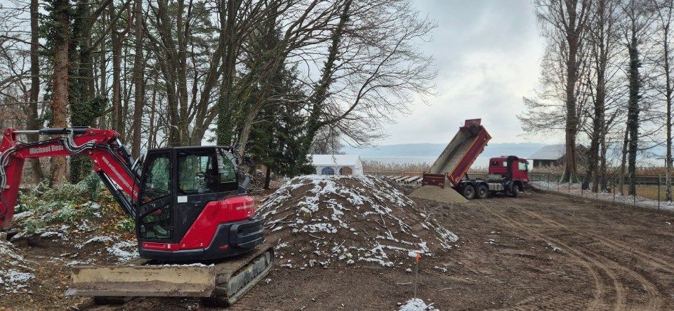 Ein roter Bagger und ein Muldenkipper sind an einem bewölkten Tag auf einer Baustelle in der Nähe von Bäumen und einem Gewässer im Einsatz.