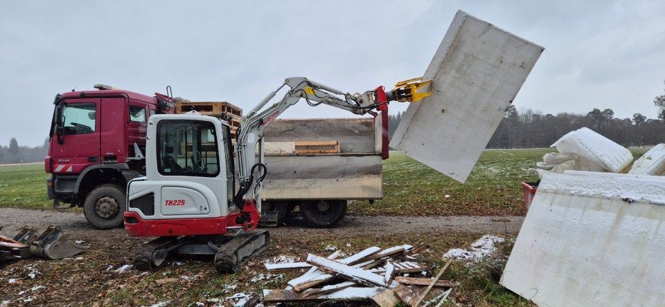 Ein Bagger verlädt eine große Betonplatte auf einen Lkw. Bauschutt liegt verstreut auf dem Boden.