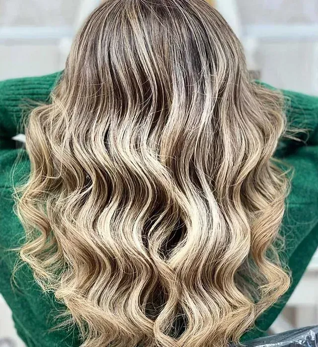 Una mujer con cabello rubio largo y ondulado lleva un suéter verde.