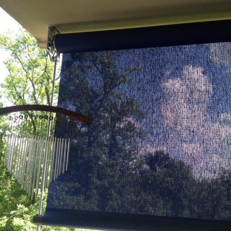 Un carillon à vent est suspendu à côté d'un store enrouleur bleu foncé sur une véranda, avec vue sur des arbres verdoyants et le ciel au loin.