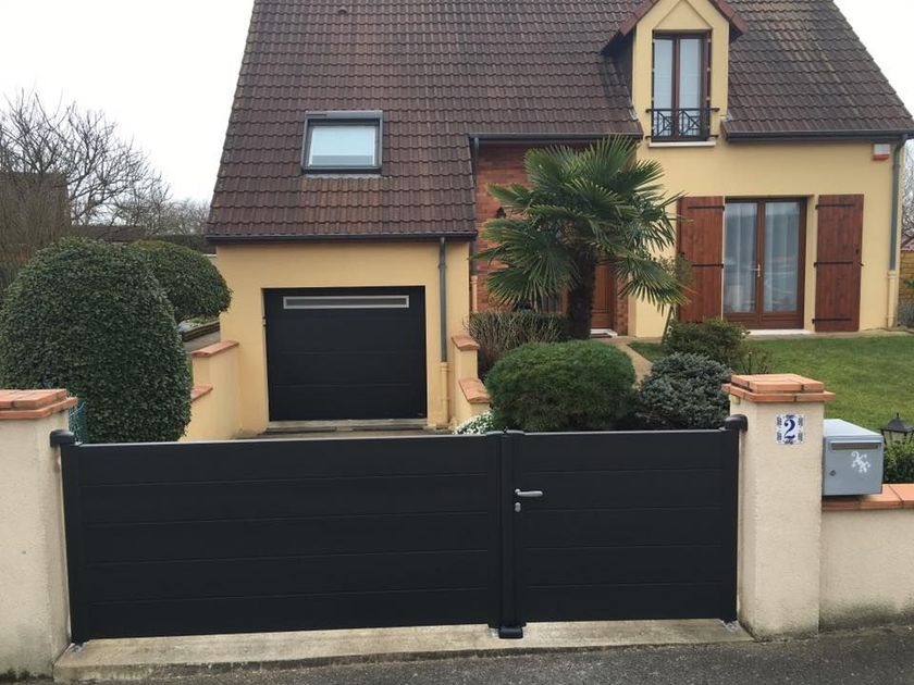 Une maison à deux étages aux murs beiges et au toit marron, derrière un portail d'allée gris foncé et des piliers couleur crème.