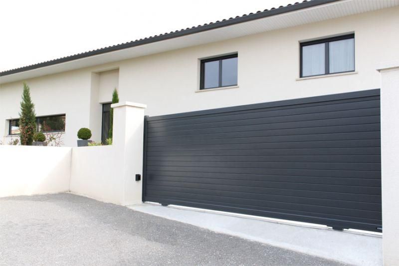 Un portail coulissant moderne à lames horizontales grises, installé devant une maison blanche aux encadrements de fenêtres noirs.