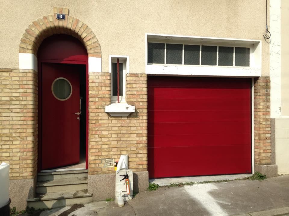 Une porte rouge ouverte avec une fenêtre ronde et une porte de garage rouge fermée sur la façade d'un bâtiment en briques beiges.