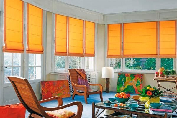 Une pièce baignée de soleil avec des stores romains orange, des chaises en bois, de l'art abstrait et une table basse en verre avec une coupe de fruits.