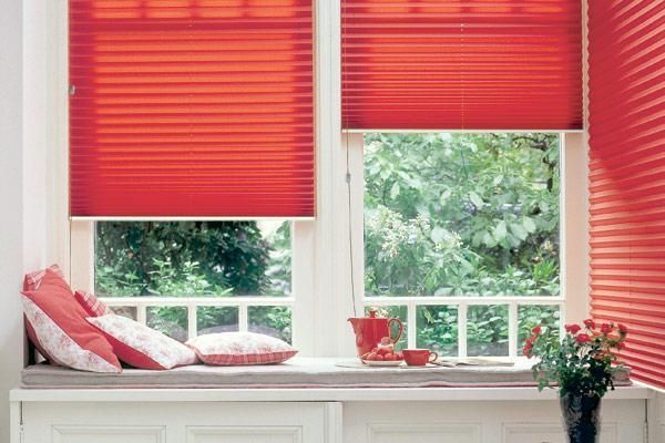 Des stores plissés rouge vif recouvrent partiellement deux fenêtres au-dessus d'une banquette rembourrée ornée de coussins à motifs et de fleurs.