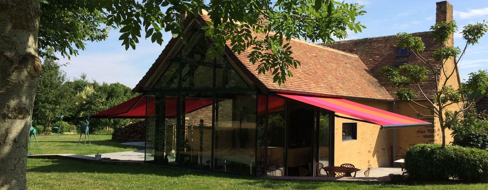 Un cottage historique avec une extension moderne en verre et des auvents rouges, situé dans un jardin ensoleillé et verdoyant.