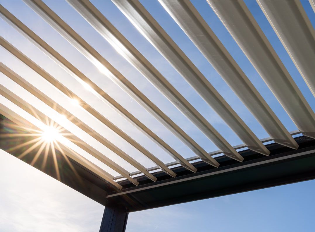 La lumière du soleil filtre à travers les lattes réglables d'une pergola extérieure moderne, sur un ciel bleu limpide.