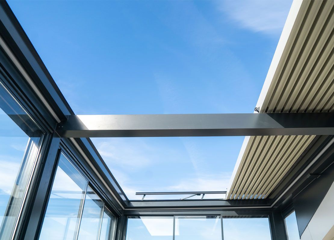 Vue en contre-plongée d'une véranda ou d'un toit en verre moderne, montrant des poutres structurelles sombres se détachant sur un ciel bleu clair.