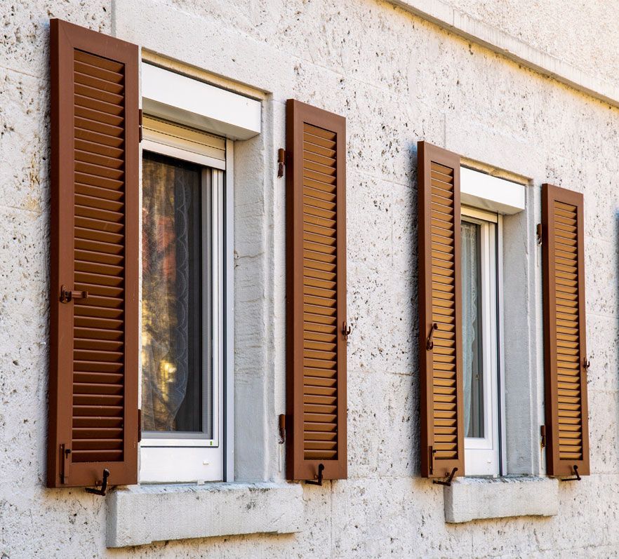 Des volets persiennes marron sont fixés à la façade d'un bâtiment en pierre blanche texturée, de part et d'autre de deux fenêtres.