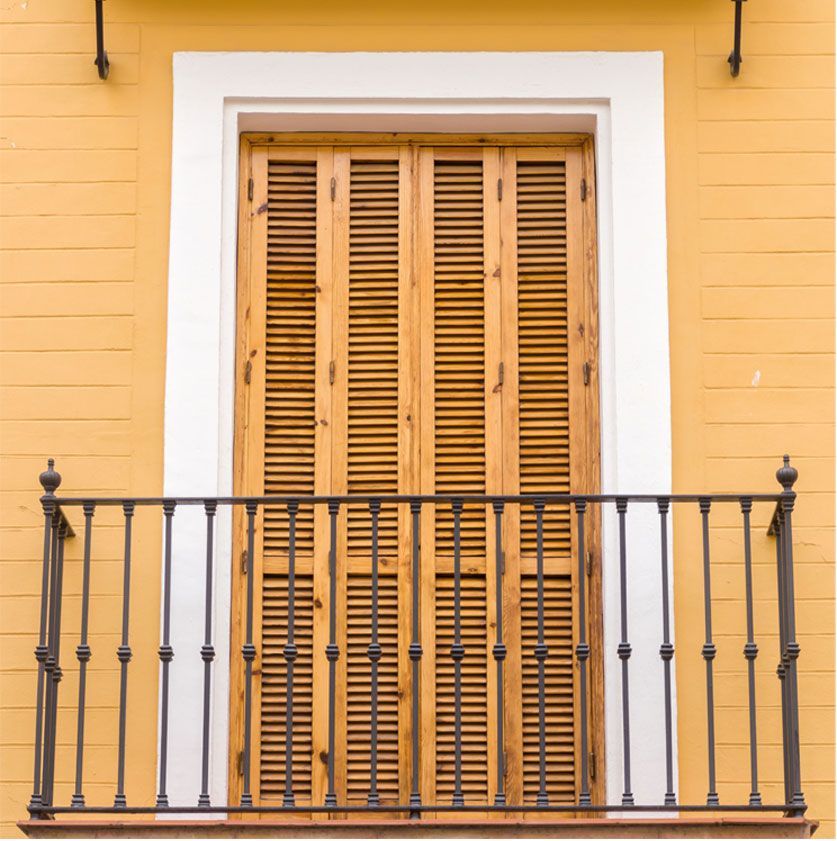 Une fenêtre en bois à persiennes fermées, encadrée de blanc, se détache sur un mur jaune avec une rambarde de balcon en métal noir.
