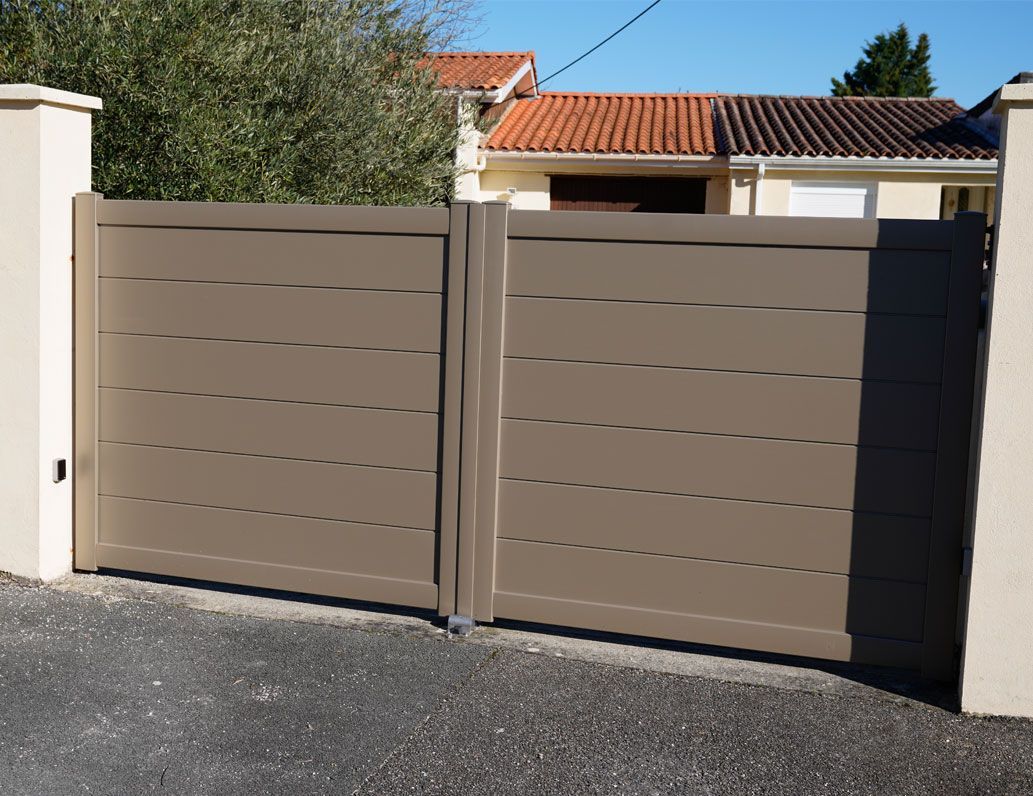Un portail battant double moderne, de couleur taupe, à lattes horizontales, entre deux piliers de pierre, installé dans une allée résidentielle.