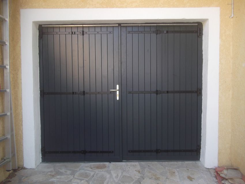 Un ensemble de portes doubles à panneaux verticaux gris foncé, avec des charnières en métal noir, encadrées de blanc, sur une terrasse en pierre.