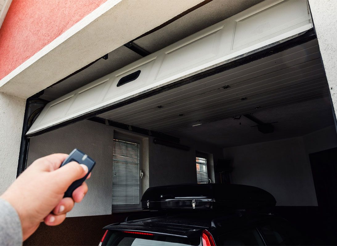 Une main tenant une télécommande pointée vers une porte de garage entrouverte, une voiture étant garée à l'intérieur.