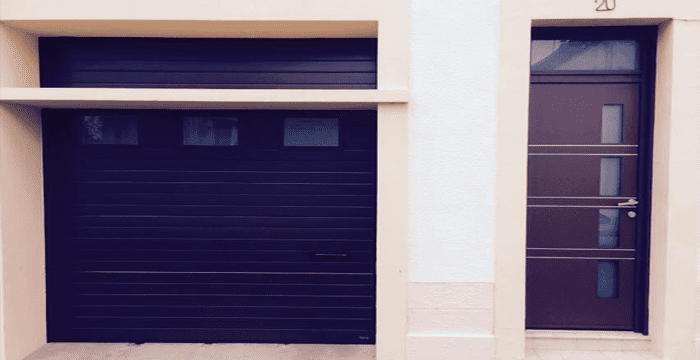Une porte de garage brun foncé, dotée de trois petites fenêtres, côtoie une porte d'entrée moderne assortie, avec des panneaux de verre verticaux.