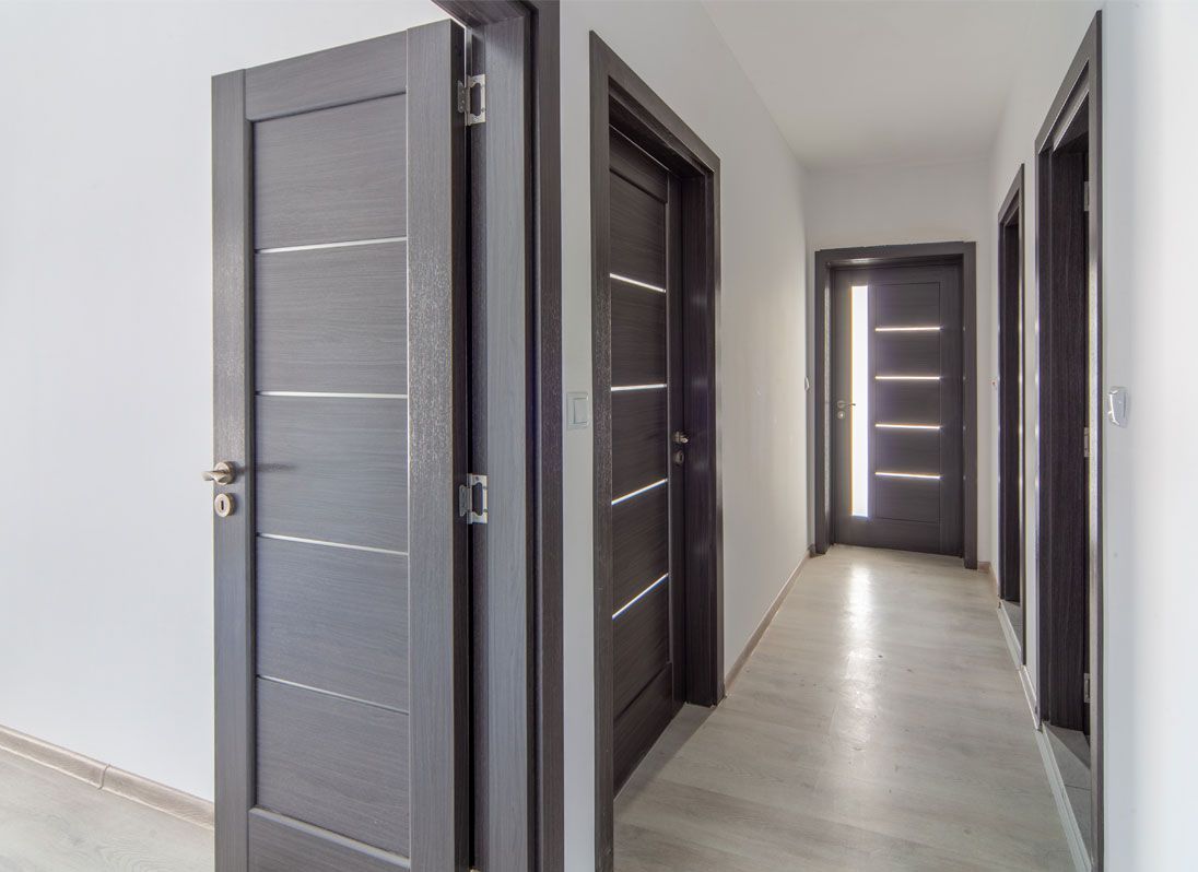 Un couloir aux portes en bois foncé ornées d'incrustations métalliques horizontales, se détachant sur des murs blancs et un sol clair.