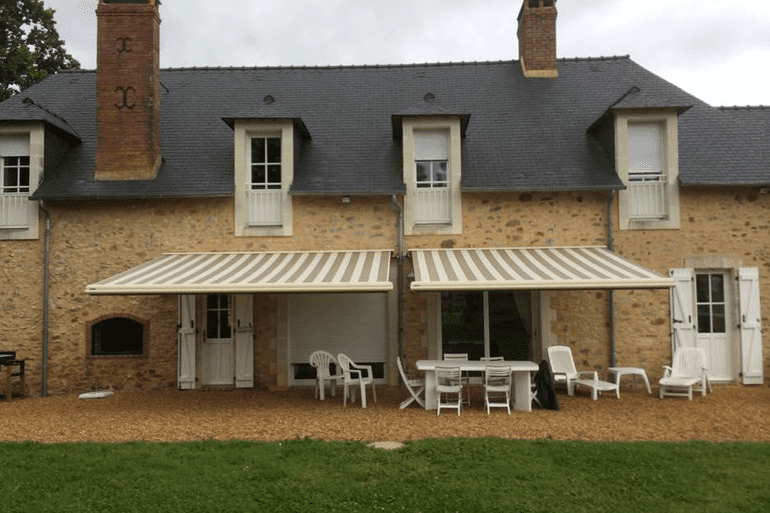Une maison en pierre avec des auvents rétractables à rayures au-dessus d'une terrasse avec mobilier de jardin et chaises sur une zone gravillonnée.