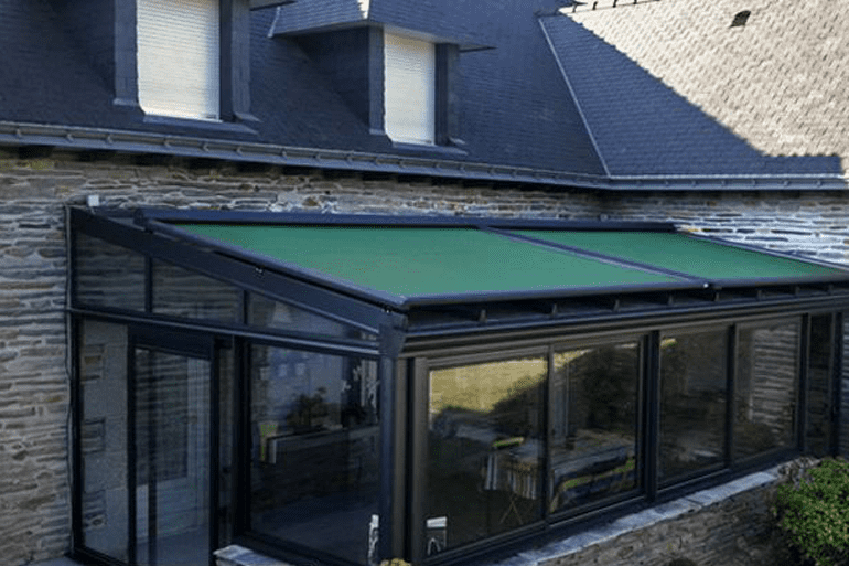 Une véranda vitrée avec un toit rétractable vert, attenante à une maison en pierre.