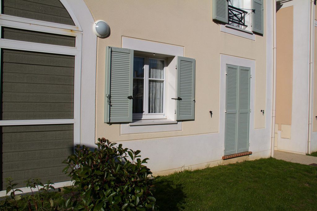 Un bâtiment à l'extérieur beige, avec une fenêtre à volets verts et une porte assortie, se détachant sur une petite pelouse verte.