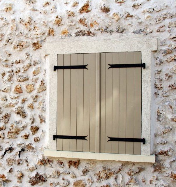 Un mur de pierre entoure une fenêtre carrée recouverte de deux volets en bois beige clair à charnières en fer noir.