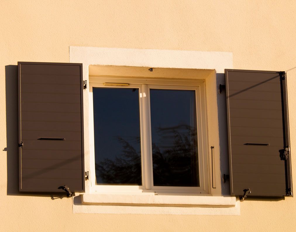 L'extérieur d'un bâtiment de couleur claire présente une fenêtre avec des volets brun foncé ouverts de chaque côté.